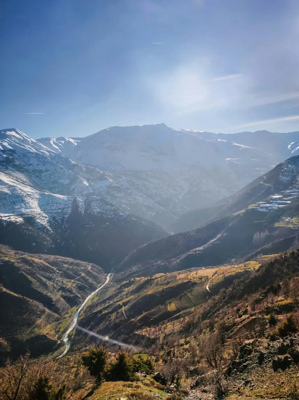 The image shows a valley with snow-capped mountains in the background. In the foreground, there is a river running through the valley. There are trees and bushes on either side of the river. The sky is blue and there are a few clouds in the sky. The image is taken from a high vantage point, looking down into the valley. The mountains in the background are tall and imposing, and the snow-capped peaks add to their beauty. The river in the foreground is winding and meandering, and the trees and bushes on either side provide a sense of lushness and greenery.