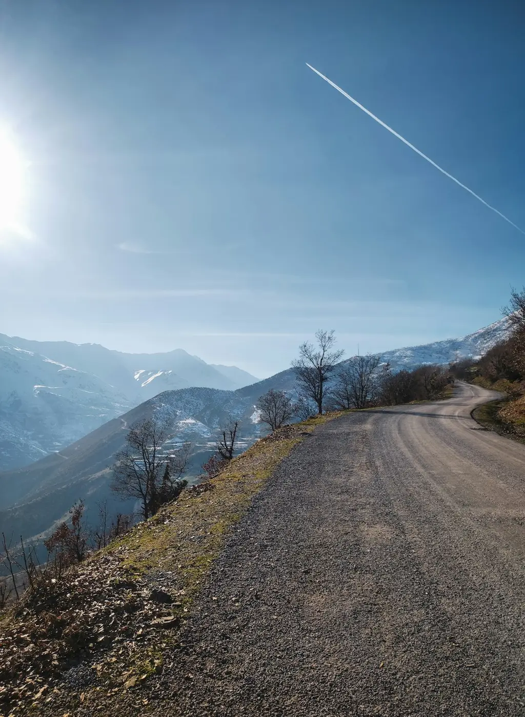 The image shows a dirt path winding through snow-capped mountains. There are pine trees on either side of the path. The sky is clear and blue.