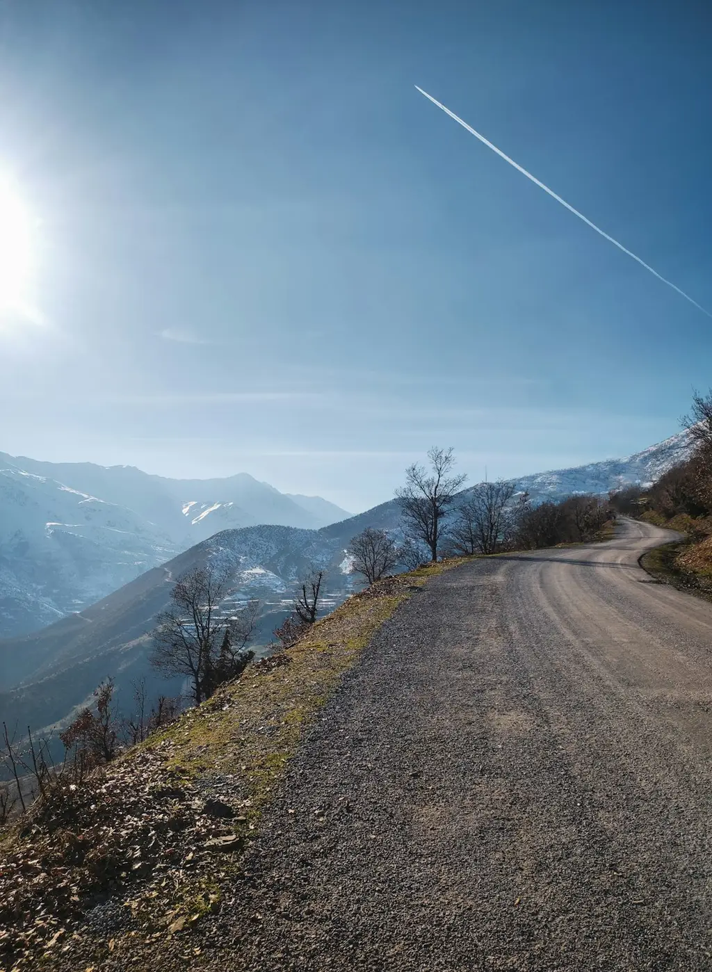 The image shows a dirt path winding through snow-capped mountains. There are pine trees on either side of the path. The sky is clear and blue.